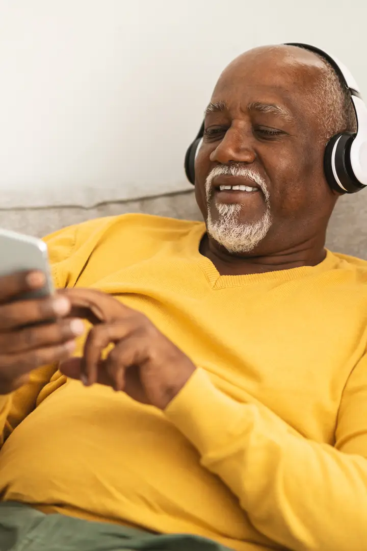 A smiling older man in headphones relaxes on his couch, choosing music on his phone