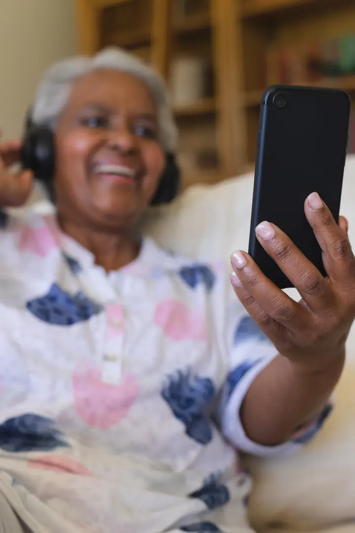 A laughing older woman in headphones holds up her phone on the sofa